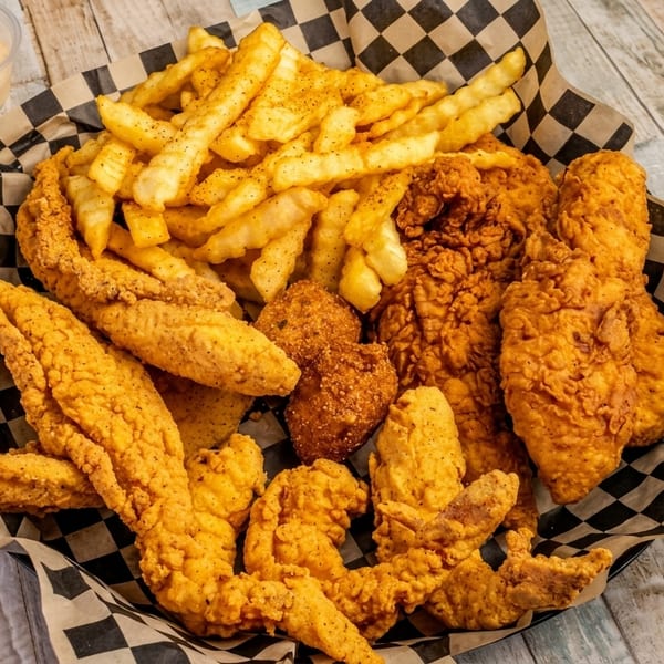 A basket of fried chicken, fish, hush puppies, and french fries with a side of dipping sauce.