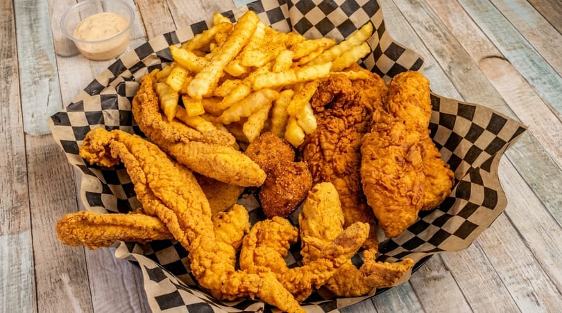 A basket of crispy fried chicken, fish, hush puppies, and french fries with a side of sauce.