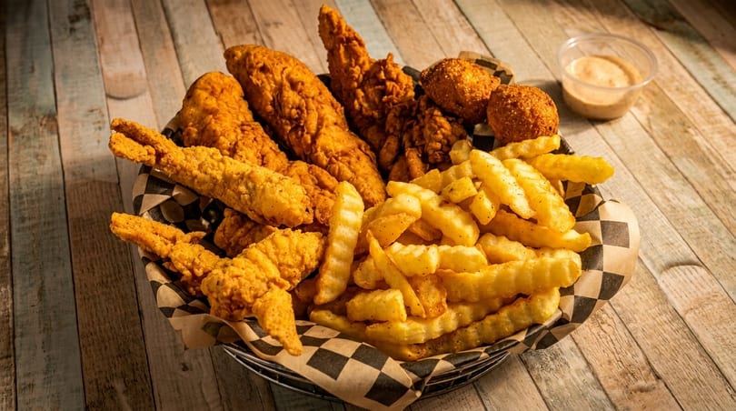 A basket of crispy fried chicken tenders, hush puppies, and crinkle-cut fries with dipping sauce.