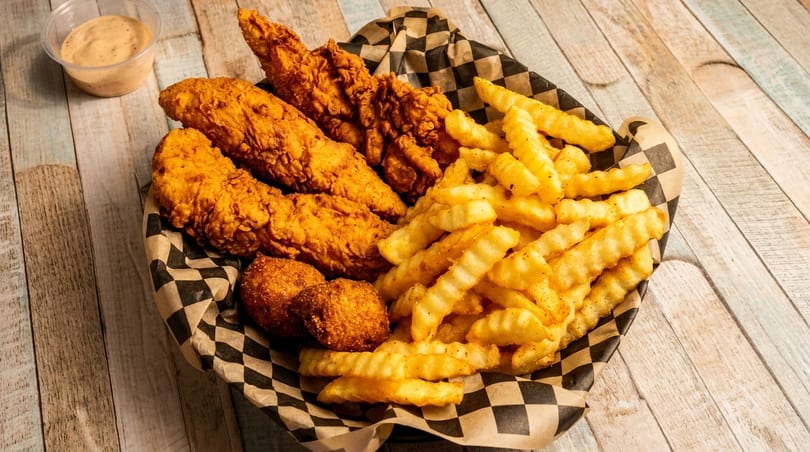 A basket of crispy fried chicken tenders, hush puppies, and crinkle-cut fries with dipping sauce.